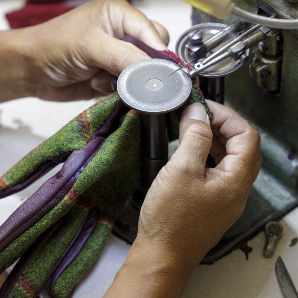Artisan hands guiding a sewing machine as they attach a vibrant green and purple leather glove, showcasing Italian craftsmanship.